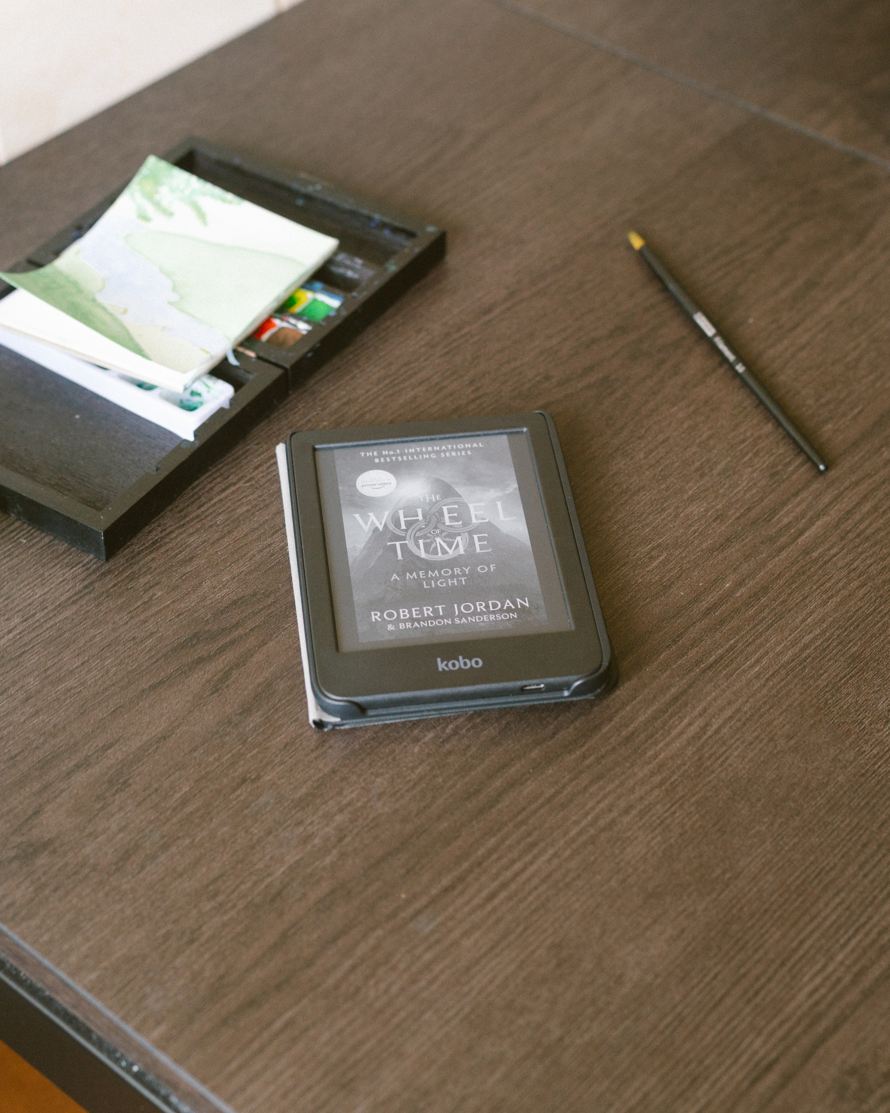 Auto-generated description: A Kobo e-reader displaying The Wheel of Time by Robert Jordan is placed on a wooden table alongside a pen and a black tray with papers.