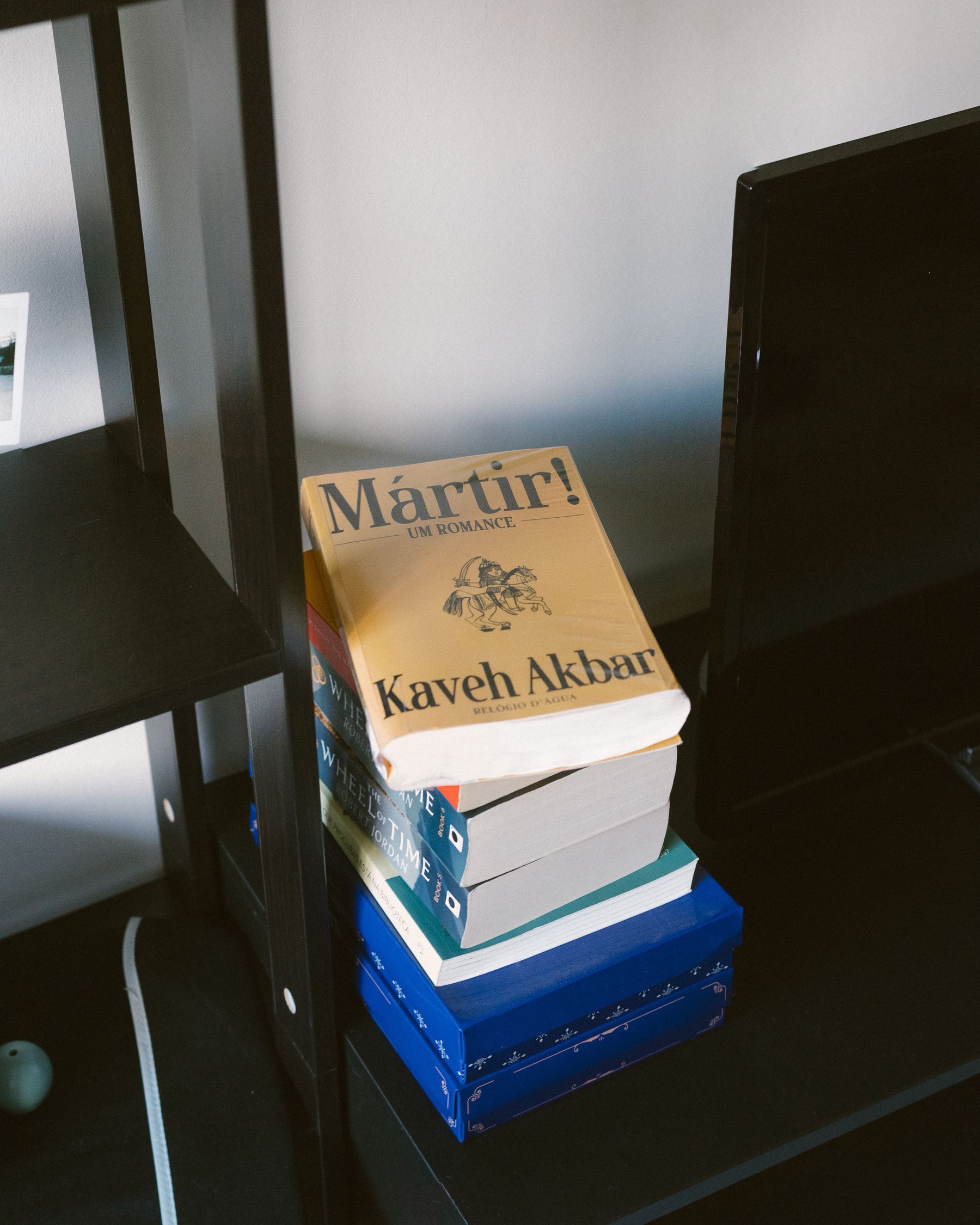 Auto-generated description: A stack of books, topped with Märtír! by Kaveh Akbar, is placed on a black shelf next to a television.